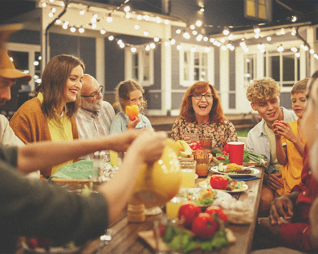 A family enjoying an outdoor dinner with a home visible in the background and string lights overhead