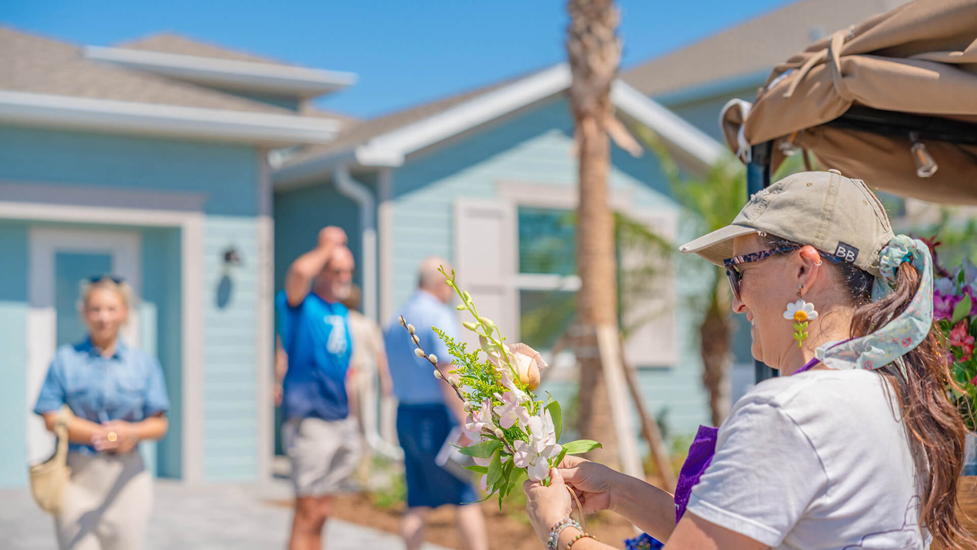 A woman in the foreground, holding a bunch of flowers, is enjoying the reveal of New Southwest Florida Homes by Lennar, alongside a small group of smiling people on a sunny Florida day