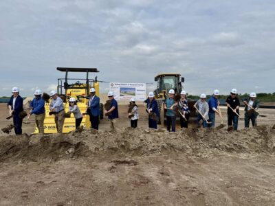 a row of men and women in hardhats turn earth with shovels in hand, all in front of large construction equipment, as part of the ground breaking ceremony for Rye Ranch Elementary School in Parrish Florida