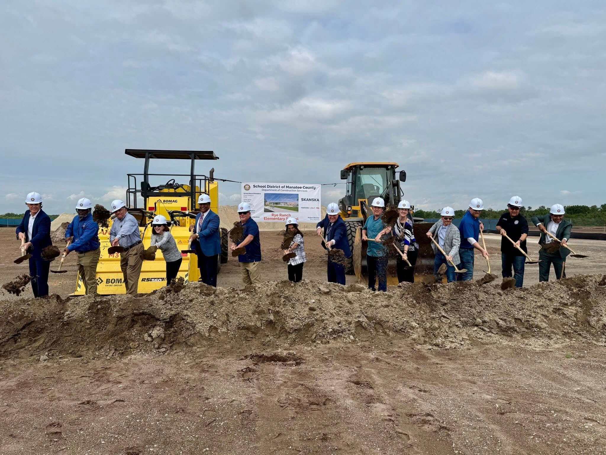 a row of men and women in hardhats turn earth with shovels in hand, all in front of large construction equipment, as part of the ground breaking ceremony for Rye Ranch Elementary School in Parrish Florida