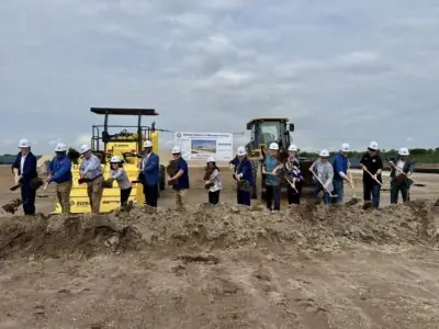 a row of men and women in hardhats turn earth with shovels in hand, all in front of large construction equipment, as part of the ground breaking ceremony for Rye Ranch Elementary School in Parrish Florida