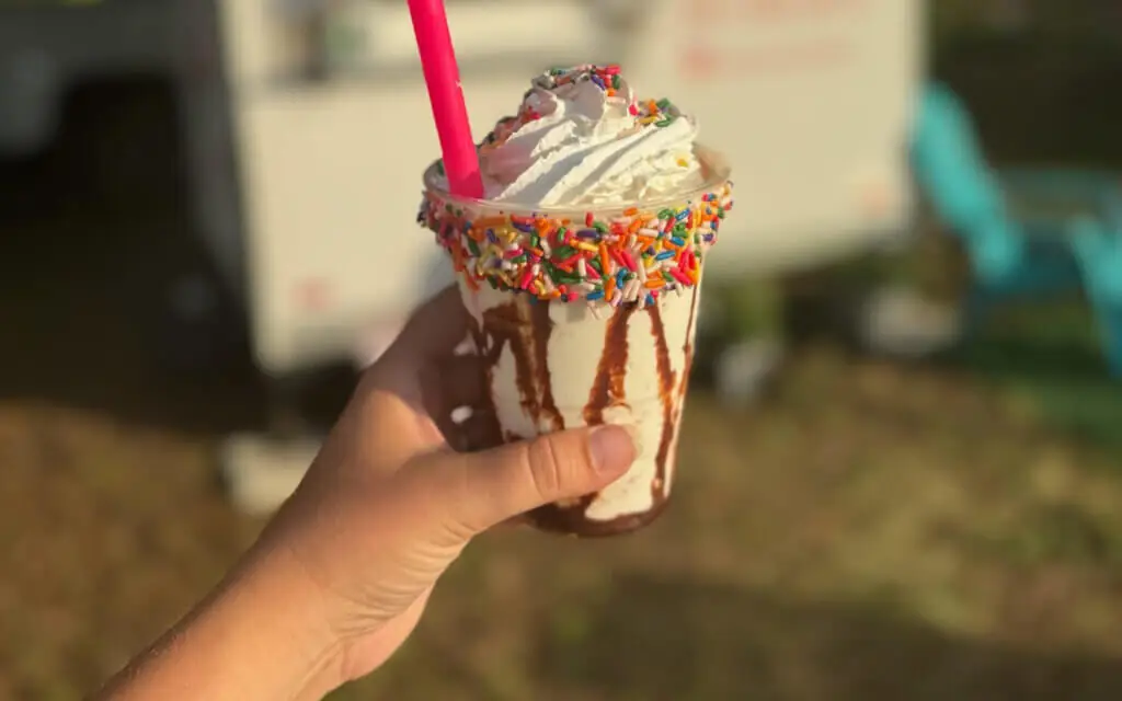 a milkshake with a chocolate swirl and topped by whipped cream and colorful sprinkles is held aloft with Sunsets & Sweets in soft focus in the background